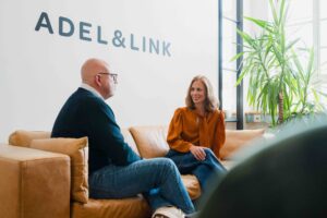 Matthias Adel und Natalie Link sitzen im neuen Büro auf dem Sofa unter dem Schriftzug von Adel & Link