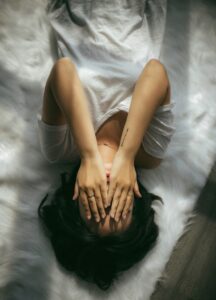 Dunkelhaarige Frau von oben fotografiert in einem Bett liegend mit den Händen vor dem Gesicht. Symbolisiert Müdigkeit und Erschöpfung.