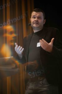 Das Foto zeigt Matthias Metzler in schwarzem Anzug vor einem dunklen Hintergrund, sprechend und gestikulierend.