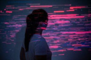 Zu sehen ist eine langhaarige Frau mit Zopf von hinten, die angestrahlt wird von pinkfarbenen Lichtreflexen, die bei genauerem Hinsehen Datenzeilen auf einem Computer sind. Symbol für Verlorensein im Web.