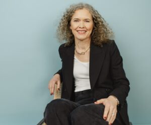 Anke Sinnigen, Autorin und Gründerin von wexxeljahre.de, lächelt in die Kamera, sitzend vor petrolfarbenem Hintergrund.
