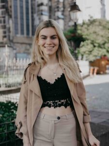 Autorin Antonia Wesseling in schwarzem Top und beigefarbenem Kostüm auf sonnenbeschienener Straße. Die blonde junge Frau lächelt offen in die Kamera.