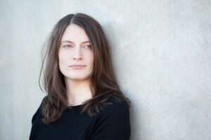 Die braunhaarige Autorin Lena Kiefer ist zu sehen vor einer weißen Wand mit in die Ferne schweifendem Blick.
