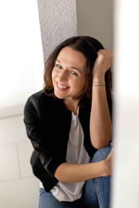 Zu sehen ist Toni Winter, Autorin, sie streicht sich durchs Haar, während sie in die Kamera lächelt, trägt Jeans und weißes Shirt mit schwarzem Jäckchen.