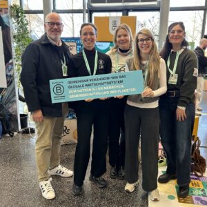 Eine Gruppe von 5 Personen, 1 Mann, 4 Frauen, auf dem BCorp-Impact-Festival. In der Hand halten sie ein Schild, auf dem steht: Gemeinsam verändern wir das globale Wirtschaftssystem zum Nutzen aller Menschen, Gemeinschaften und des Planeten.