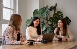 Interviewsituation mit drei lächelnden Frauen an einem Tisch sitzend, drei Kaffeetassen mit Adel & Link Logo und ein Laptop auf dem Tisch. Grünpflanze im Hintergrund.