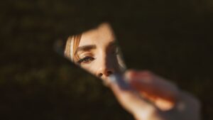 Hand hält Spiegelscherbe mit gespiegeltem Gesicht einer Frau vor dunklem Hintergrund